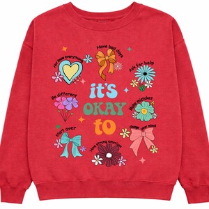 May include: Red sweatshirt with the phrase "it's OKAY to" and various positive affirmations and illustrations, including flowers, bows, and a heart. The sweatshirt has a crew neck and long sleeves.