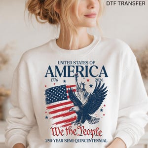 May include: White sweatshirt featuring a patriotic design. The graphic includes the American flag, an eagle, and the Statue of Liberty. Text reads "UNITED STATES OF AMERICA", "We the People", and "250-YEAR SEMI-QUINCENTENNIAL".