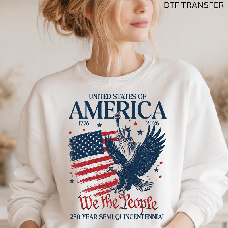 May include: White sweatshirt featuring a patriotic design. The graphic includes the American flag, an eagle, and the Statue of Liberty. Text reads "UNITED STATES OF AMERICA", "We the People", and "250-YEAR SEMI-QUINCENTENNIAL".