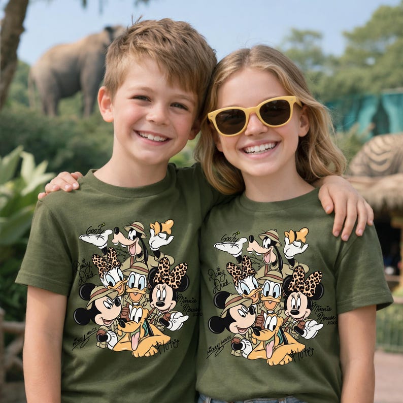 Puede incluir: Dos ni&ntilde;os vistiendo camisetas verde oliva con un dise&ntilde;o de dibujos animados de personajes de Disney con atuendos de safari. El dise&ntilde;o incluye Mickey Mouse, Minnie Mouse, Donald Duck, Daisy Duck, Goofy y Pluto. El texto "Goofy", "Minnie Mouse" y "Mickey Mouse" tambi&eacute;n es visible.