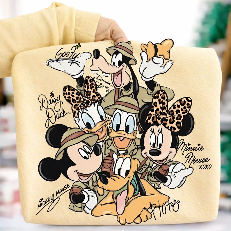 Puede incluir: Un bolso tote amarillo claro con personajes de Disney con atuendos de safari. Mickey Mouse, Minnie Mouse, Donald Duck, Daisy Duck, Goofy y Pluto est&aacute;n representados. Los nombres de los personajes y "XOXO" tambi&eacute;n est&aacute;n presentes.
