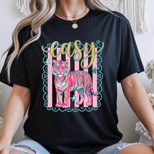 May include: Black t-shirt featuring a colorful tiger illustration with the word "easy" in a decorative font. The tiger is depicted in shades of pink, blue, and yellow, set against a pink and white striped background with star accents. The shirt is a casual wear item.