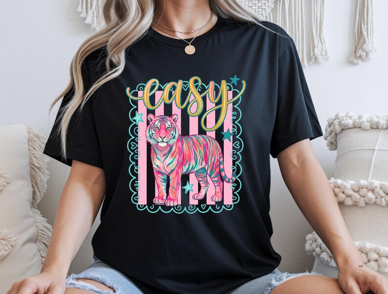 May include: Black t-shirt featuring a colorful tiger illustration with the word "easy" in a decorative font. The tiger is depicted in shades of pink, blue, and yellow, set against a pink and white striped background with star accents. The shirt is a casual wear item.