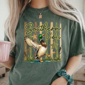 May include: Green t-shirt with the words "Southern Vibes" and a graphic of a mallard duck with outstretched wings. The design includes stars, cattails, and a water scene. The shirt has a relaxed fit.