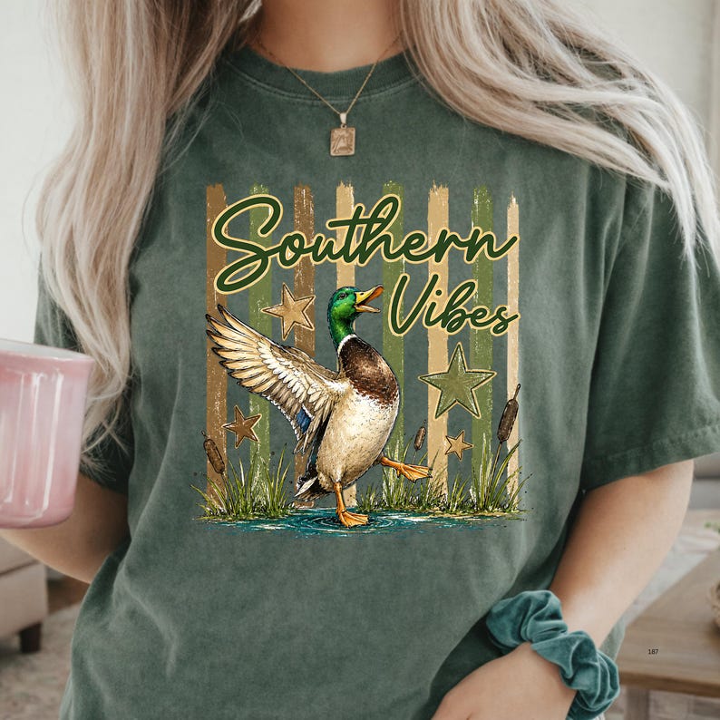 May include: Green t-shirt with the words "Southern Vibes" and a graphic of a mallard duck with outstretched wings. The design includes stars, cattails, and a water scene. The shirt has a relaxed fit.