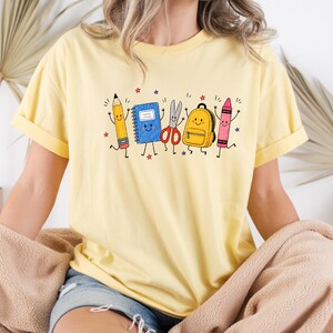 May include: A light yellow t-shirt featuring a graphic print of anthropomorphic school supplies including a yellow pencil, a blue notebook, red scissors, a yellow backpack, and a pink crayon, all with smiling faces and thin black limbs.