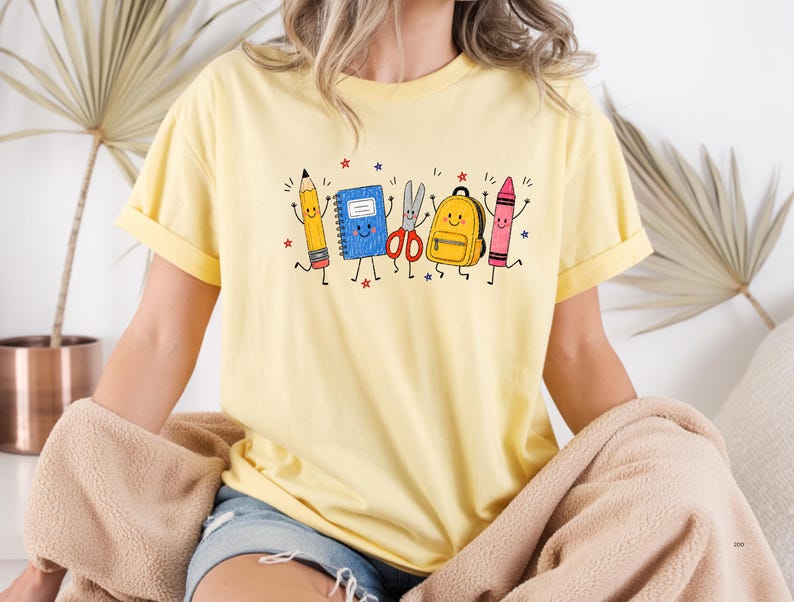 May include: A light yellow t-shirt featuring a graphic print of anthropomorphic school supplies including a yellow pencil, a blue notebook, red scissors, a yellow backpack, and a pink crayon, all with smiling faces and thin black limbs.