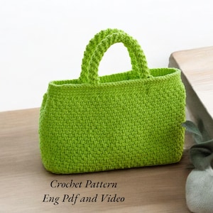 May include: A bright green crocheted tote bag with a thick, crocheted handle. The bag is sitting on a wooden surface. The text "Crochet Pattern Eng Pdf and Video" is below the bag.