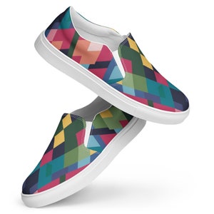 Op de afbeelding: Een paar witte canvas slip-on schoenen met een kleurrijk geometrisch patroon. Het patroon bevat tinten rood, blauw, groen, geel en zwart.