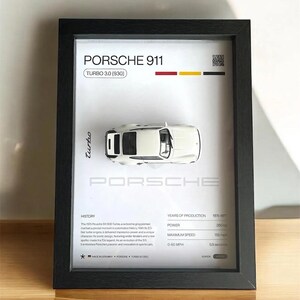 Puede incluir: Impresión enmarcada con un modelo de coche Porsche 911 Turbo 3.0 (930) blanco. La impresión incluye el nombre del coche, especificaciones y una breve historia. El marco es negro y el fondo blanco.