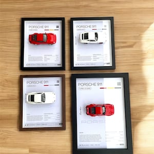 Peut inclure: Quatre impressions encadrées de différents modèles de Porsche 911. Chaque impression présente un fond blanc avec du texte noir et une petite voiture miniature au centre. Les modèles sont une Porsche 911 Turbo 3.0 (930) rouge, une Porsche 911 Carrera RS 2.7 blanche, une Porsche 911 Carrera 3.2 blanche et une Porsche 911 Turbo 3.3 rouge.