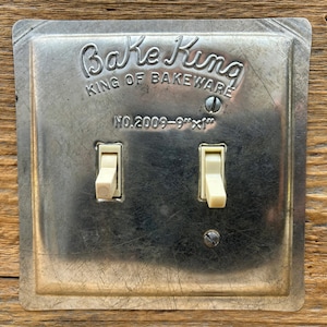 May include: Vintage Bake King light switch plate with two ivory-colored toggle switches. The metal plate is square with rounded corners and embossed text that reads "Bake King, King of Bakeware". The plate measures 9" x 1".