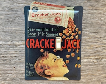 May include: A rectangular light switch cover featuring a vintage Cracker Jack advertisement. The design includes a child looking up at falling popcorn, the text Gee-wouldn't it be Great if it Snowed, Cracker Jack, and The More You Eat The More You Want.