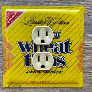 May include: A custom electrical outlet cover featuring two white outlets set against a yellow background. The background is a Nabisco Wheat Thins snack cracker box with the words "Limited Edition" and "Wheat Thins" printed on it. The net weight is 13 oz.