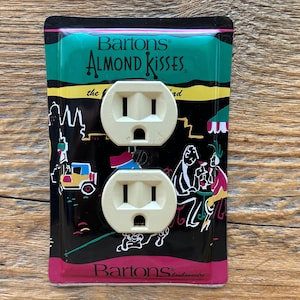 May include: A vintage electrical outlet cover featuring two beige outlets. The cover has a black background with colorful illustrations and the text "Bartons Almond Kisses". The design includes a cityscape and figures at a table.