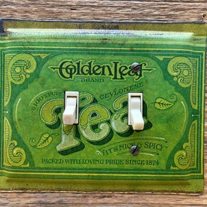 May include: Vintage green Golden Leaf tea light switch cover with two white switches. The cover features the words "Golden Leaf Brand Ceylon Tea" and "It's Nice & Spicy" with the text "Packed with Loving Pride Since 1874".