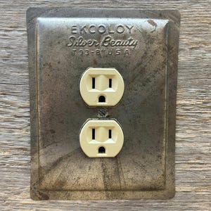 May include: Vintage EKCOLOY Silver Beauty electrical outlet cover with two cream-colored outlets. The metal cover has a distressed, aged appearance with the text "EKCOLOY Silver Beauty T93-B USA" embossed on the surface.