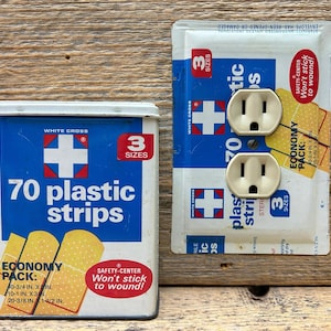 May include: A vintage first aid kit with a blue and white tin containing 70 plastic strips. The tin has a red cross logo and the text "3 sizes". Next to it is a wall outlet cover with a matching design and two electrical outlets.