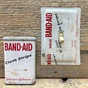 May include: A vintage rectangular metal Band-Aid tin for cloth strips and a matching light switch cover plate. Both items feature the red and white Band-Aid branding, the text Cloth Strips, and the Johnson & Johnson logo on a white background.