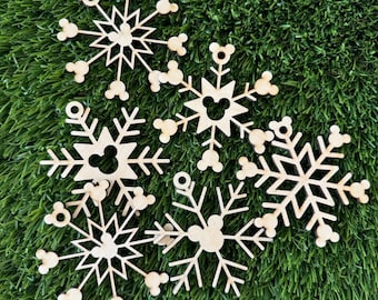 May include: Six wooden snowflake ornaments with a Mickey Mouse head cutout in the center of each snowflake. The snowflakes are on a green artificial grass background.