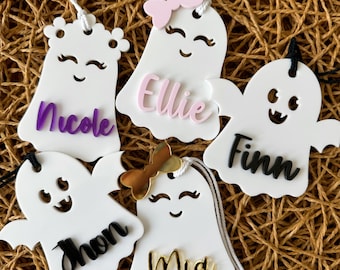 May include: Five white ghost-shaped ornaments with different names written on them. The names are Nicole, Ellie, Finn, Jhon, and Mia. Each ghost has a different facial expression and some have bows.