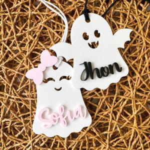 Op de afbeelding: Twee witte, spookvormige ornamenten met roze en zwarte details. Het spook links heeft een roze strik en de naam "Sofia" in roze geschreven. Het spook rechts heeft de naam "Jhon" in zwart geschreven.