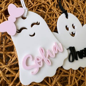 May include: Two white ghost-shaped ornaments with faces and names. The first ghost has a pink bow and the name "Sofia" written in pink. The second ghost has a black name tag with the name "Drew" written in black.