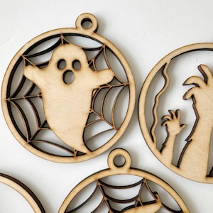 Op de afbeelding: Drie onafgewerkte houten Halloween-versieringen. Een versiering heeft een geest in een spinnenweb, een andere heeft een griezelige hand en de derde heeft een spinnenweb.