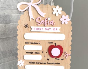 Puede incluir: Cartel de madera para el primer día de escuela con el nombre "Sofia" en escritura rosa. El cartel incluye espacios para escribir detalles sobre el primer día del niño, con lazos y flores.