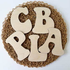 May include: Five unfinished wooden letters spelling out the word "PLAY" in a whimsical font. The letters are arranged in a circle on a woven brown background.