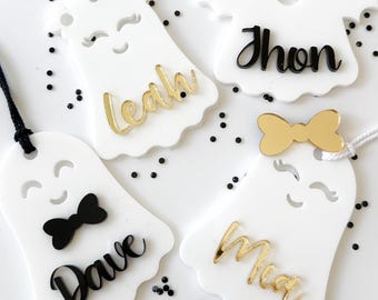 May include: Four white ghost-shaped ornaments with black and gold accents. Each ghost has a different name written on it: Leah, Jhon, Dave, and Mia.