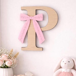 Könnte beinhalten: Buchstabe "P" aus Holz mit einer rosa Schleife. Der Buchstabe ist aus hellem Holz gefertigt und an einer weißen Wand befestigt. Die rosa Schleife hat ein Blumenmuster. Das Bild deutet auf eine Kinderzimmerdekoration hin.