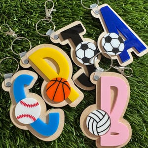 Peut inclure: Porte-clés avec des lettres et des ballons de sport. Les lettres sont bleues, jaunes et roses, avec des ballons de sport dont un de baseball, de basket-ball, de football et de volley-ball. Chaque porte-clés a un fermoir argenté.