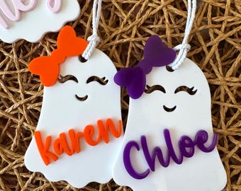 May include: White ghost-shaped name tags with orange and purple bows. The tags have smiling faces and the names "Karen" and "Chloe" in orange and purple script. Each tag has a white string for hanging.