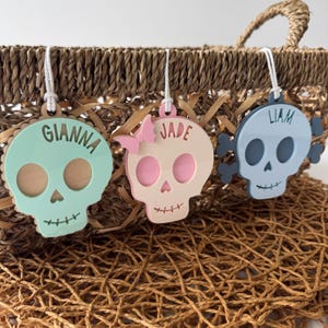 Op de afbeelding: Drie decoratieve schedelvormige ornamenten met namen, hangend aan een gevlochten mand. Eén is mintgroen met "GIANNA", een andere is roze met "JADE" en een vlinder, en de laatste is lichtblauw met "LIAM" en kruisbeenderen.