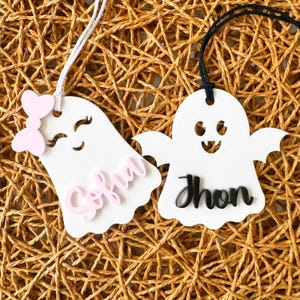 Op de afbeelding: Twee witte, spookvormige ornamenten met zwarte en roze letters. Het spook links heeft een roze strik en de naam "Sofia" in roze geschreven. Het spook rechts heeft de naam "Jhon" in zwart geschreven.
