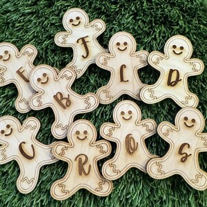 May include: Wooden gingerbread man ornaments with letters etched on them. The ornaments are light brown with a smiling face and a decorative outline. The letters include F, B, C, R, O, S, J, L, and D. The ornaments are on a green surface.