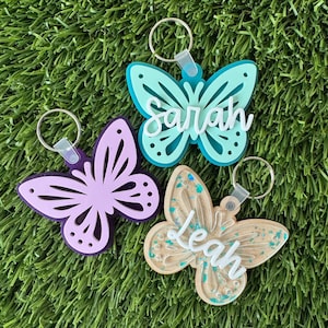 May include: Three butterfly-shaped keychains in shades of purple, teal, and tan, each with a name cut out in white. The teal keychain reads "Sarah", the tan keychain reads "Leah". The keychains are on a green surface.