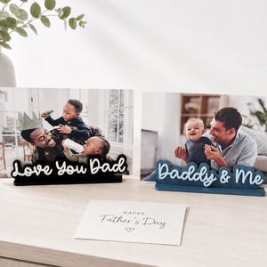 May include: Two photo frames with family photos and decorative text. One frame has the text "Love You Dad" in black, and the other says "Daddy & Me" in blue. A Father's Day card is also present.
