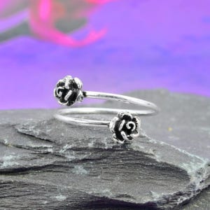 Könnte beinhalten: Ein silberfarbener Ring mit zwei Rosenblüten-Designs. Der Ring umschlingt den Finger mit einer Rose an jedem Ende. Die Rosen haben detaillierte Blütenblätter und sind leicht oxidiert. Der Ring wird auf einer dunkelgrauen, strukturierten Oberfläche präsentiert.