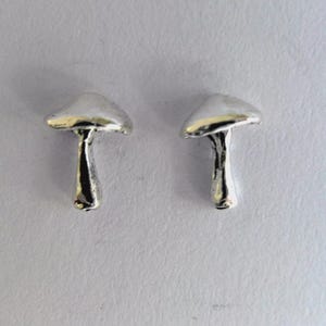 May include: Two small, silver mushroom charms. Each charm has a rounded cap and a slender stem. The mushrooms are displayed against a plain, light-coloured background. These charms could be used for jewellery or other craft projects.