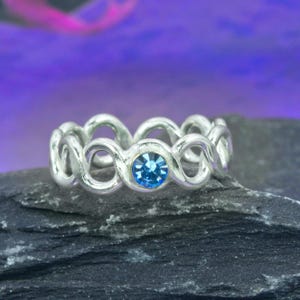 Pode incluir: Um anel prateado com um design de laços infinitos repetidos. O anel apresenta uma gema azul brilhante redonda no centro. É exibido em uma superfície texturizada cinza escuro, com um fundo roxo desfocado. Design elegante.