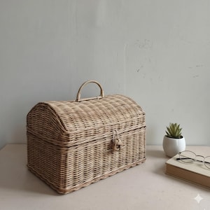 May include: A light brown woven wicker chest with a curved top and a handle. The chest has a latch closure and is sitting on a light-coloured surface. A small plant in a white pot and a book with glasses are in the background.