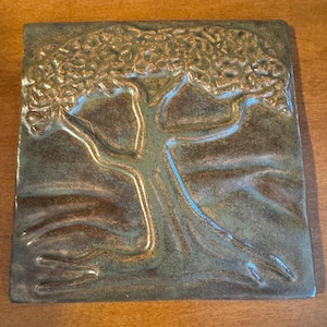 May include: A square ceramic tile featuring a raised relief design of a tree with a wide, textured canopy. The surface has a glossy, mottled glaze in shades of dark brown, bronze, and muted green, creating a dimensional effect across the tile.