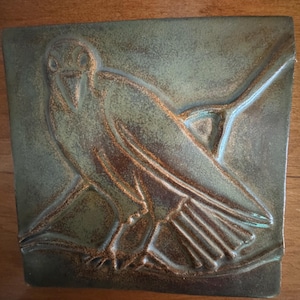 May include: A square ceramic tile featuring a relief design of a bird perched on a branch. The tile has a mottled finish with deep green and earthy brown tones, highlighting the textured details of the bird's feathers and the surrounding branch.