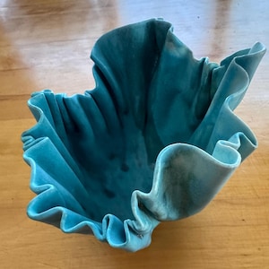 May include: A turquoise ceramic bowl with a ruffled, wavy rim and a deep, organic shape. The glaze features variations in color intensity, creating a textured appearance across the interior and exterior surfaces of the vessel.