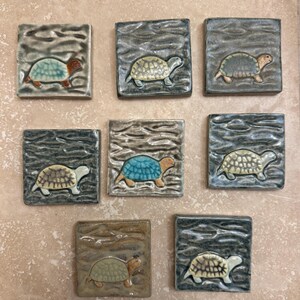 May include: A collection of eight square ceramic tiles, each featuring a raised relief of a turtle. The tiles have a textured, wavy surface in shades of gray and brown, with individual turtles painted in various colors including blue, tan, and green.