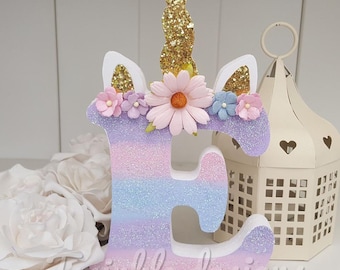 May include: A wooden letter E decorated with a unicorn horn, glitter, and pastel flowers. The letter is painted in a gradient of purple, pink, and blue.