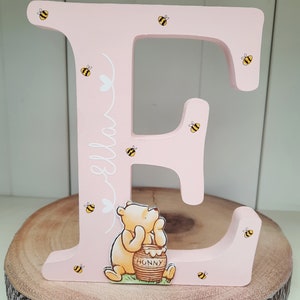 Pode incluir: Uma letra "E" de madeira rosa decorada com abelhas amarelas e pretas e uma ilustra&ccedil;&atilde;o de Winnie the Pooh. O nome "Ella" est&aacute; escrito em branco. A letra est&aacute; sobre uma fatia de madeira.