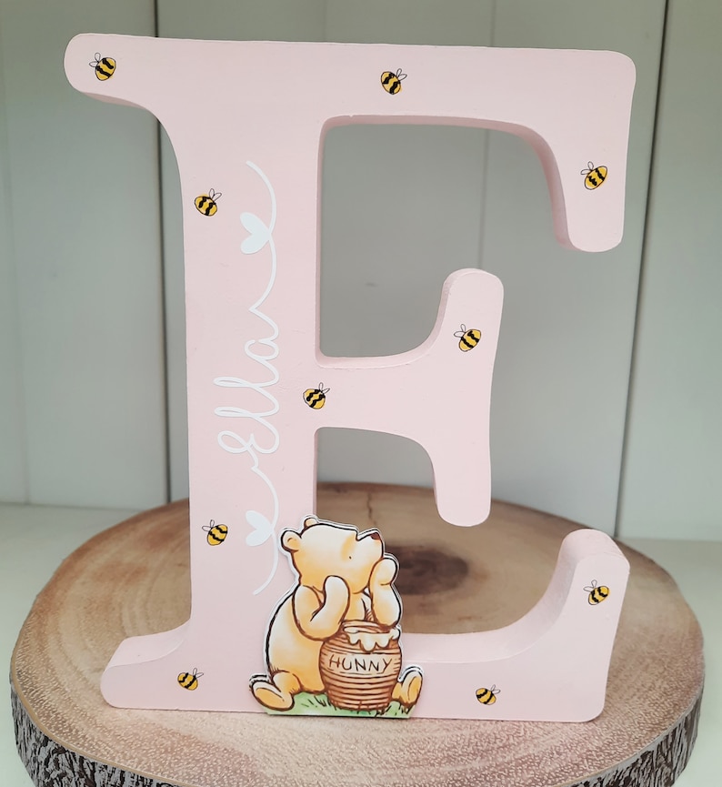 Pode incluir: Uma letra "E" de madeira rosa decorada com abelhas amarelas e pretas e uma ilustra&ccedil;&atilde;o de Winnie the Pooh. O nome "Ella" est&aacute; escrito em branco. A letra est&aacute; sobre uma fatia de madeira.